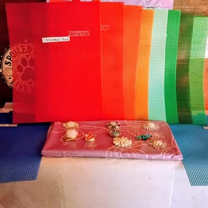 May include: A collection of rectangular plastic mesh sheets in red, orange, yellow, light green, and dark green, arranged vertically behind a mauve rectangular package featuring a white floral pattern and several assorted decorative brooches.