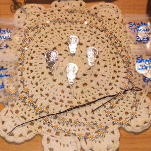May include: A daisy chain trim featuring small white and yellow flowers, four silver-toned metal skull charms, and a dark sewing needle. A white sign with the handwritten text "Tiny Yin Treasure Shore Box Sewing Notions" is visible at the top.