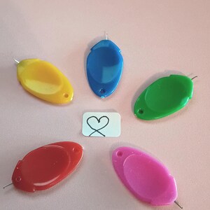 May include: Five oval-shaped plastic stitch markers in yellow, blue, green, red, and pink, arranged in a circle around a small white rectangular tag featuring a hand-drawn black heart symbol. Each marker has a small metal needle point at one end.