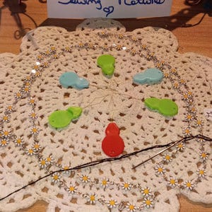May include: A collection of sewing notions including a daisy chain trim, five colorful gourd-shaped plastic needle threaders in red, green, and blue, and a metal sewing needle with black thread. A white sign reads "Ying Yin Treasure Trove Box Sewing Notions".