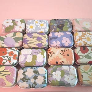May include: A collection of sixteen rectangular metal tins arranged in a grid, each featuring a unique floral pattern with various color palettes including shades of pink, blue, yellow, green, orange, and purple on a light pink background.