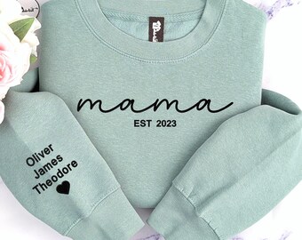 May include: A light green sweatshirt with the word "mama" in black cursive lettering and the text "EST 2023" below it. The left sleeve has the names "Oliver James Theodore" and a heart printed in black.