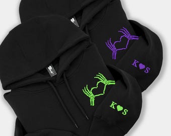Könnte beinhalten: Zwei schwarze Kapuzenpullover mit gestickten Skeletthänden, die ein Herz auf der Brust bilden. Ein Pullover hat eine grüne Stickerei, der andere eine violette. Auf beiden Ärmeln befindet sich der Text K Herz S.