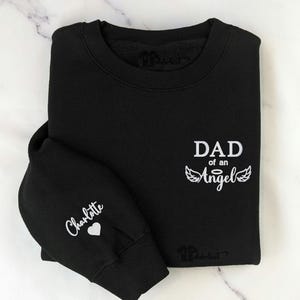 May include: Black sweatshirt with white text that reads "DAD of an Angel" with angel wings. The left sleeve has the name "Charlotte" and a heart.