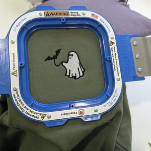 May include: A 5.5 inch blue Mighty Hoop embroidery frame securing olive green fabric. The fabric features an embroidered white ghost holding a black bat by a string. The frame includes warning text about strong magnets and electronic devices.