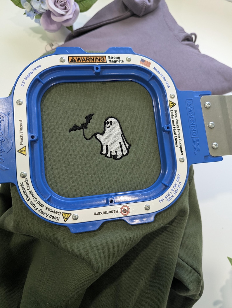 May include: A 5.5 inch blue Mighty Hoop embroidery frame securing olive green fabric. The fabric features an embroidered white ghost holding a black bat by a string. The frame includes warning text about strong magnets and electronic devices.