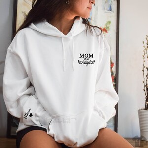May include: White hoodie with black text that reads "MOM of an Angel" and a pair of angel wings. The left sleeve has the name "Katherine" and a small black heart.