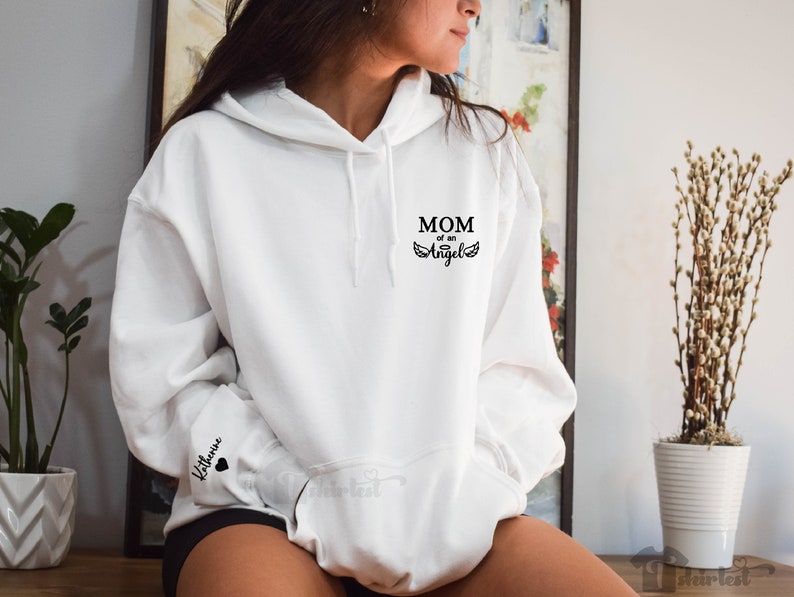 May include: White hoodie with black text that reads "MOM of an Angel" and a pair of angel wings. The left sleeve has the name "Katherine" and a small black heart.