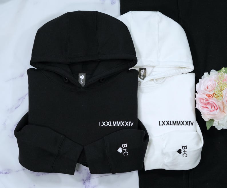 A pair of folded matching hoodies in black and white. Both feature embroidered Roman numerals on the left chest. The sleeve cuffs are customized with a small heart and the initials 'B+C' in a clean, new times Roman font, ideal for anniversary, wedding or engagement gifts.