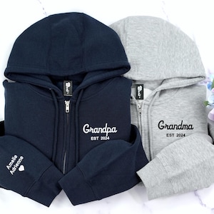 May include: Two hooded sweatshirts, one navy blue and one gray, with embroidered text. The navy blue sweatshirt has "Grandpa EST 2024" on the front and "Amelia Adrienne" on the sleeve. The gray sweatshirt has "Grandma EST 2024" on the front.