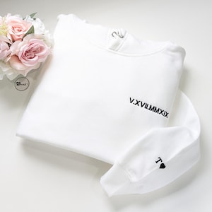 K&ouml;nnte beinhalten: Ein wei&szlig;es Sweatshirt mit dem in Schwarz gestickten r&ouml;mischen Datum "V.XVII.MMXIX". Der &Auml;rmel zeigt ein schwarzes "T" und ein Herzsymbol. Ein Strau&szlig; aus rosa Rosen und wei&szlig;en Blumen befindet sich in der oberen linken Ecke.