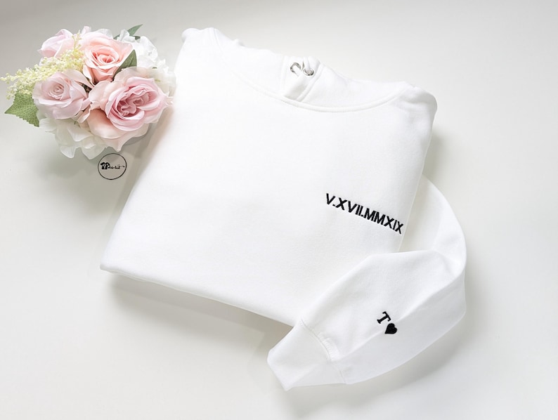 K&ouml;nnte beinhalten: Ein wei&szlig;es Sweatshirt mit dem in Schwarz gestickten r&ouml;mischen Datum "V.XVII.MMXIX". Der &Auml;rmel zeigt ein schwarzes "T" und ein Herzsymbol. Ein Strau&szlig; aus rosa Rosen und wei&szlig;en Blumen befindet sich in der oberen linken Ecke.