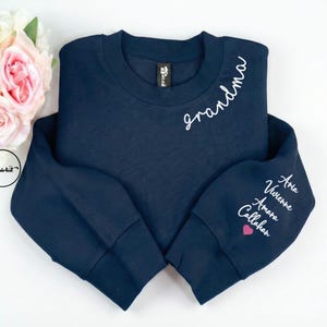 Peut inclure: Sweat-shirt bleu marine avec le mot "grandma" en écriture blanche sur la poitrine. La manche gauche porte les noms "Aria, Vivienne, Amara, Callahan" en écriture blanche, avec un cœur rose.