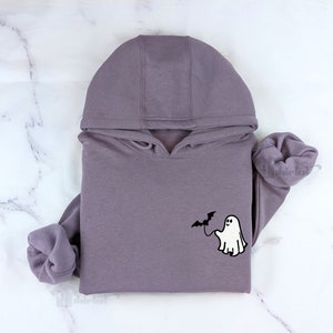 May include: A mauve hooded sweatshirt featuring a small graphic on the chest. The graphic depicts a white ghost holding a black bat on a string. The hoodie has long sleeves with ribbed cuffs and a drawstring hood, presented in a folded arrangement.
