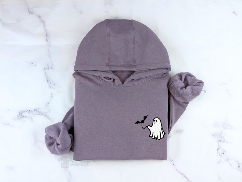 May include: A mauve hooded sweatshirt featuring a small graphic on the chest. The graphic depicts a white ghost holding a black bat on a string. The hoodie has long sleeves with ribbed cuffs and a drawstring hood, presented in a folded arrangement.