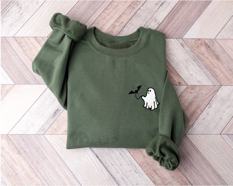 May include: An olive green crewneck sweatshirt featuring a small embroidered graphic on the left chest. The design depicts a white ghost holding a black bat on a string. The sweatshirt has long sleeves with ribbed cuffs and a ribbed neckline.