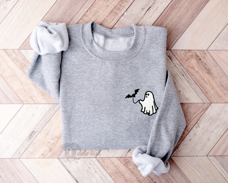 May include: A heather gray long-sleeve crewneck sweatshirt featuring a small embroidered graphic on the left chest. The design depicts a white ghost with black outlines holding a black bat on a string, creating a simple, clean aesthetic.