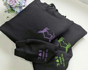 May include: Two black hooded sweatshirts featuring embroidered skeleton hands forming a heart shape on the chest. One sweatshirt has purple embroidery, the other green. The sleeves feature the text "I'm with you always" with R and K initials.