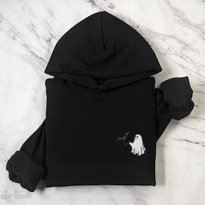 May include: A black hooded sweatshirt featuring a small graphic on the chest. The graphic depicts a white ghost holding a black bat on a string. The hoodie has long sleeves with ribbed cuffs and a hood with a drawstring opening at the top.