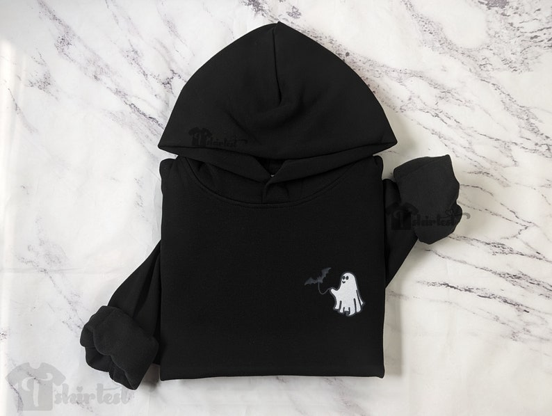 May include: A black hooded sweatshirt featuring a small graphic on the chest. The graphic depicts a white ghost holding a black bat on a string. The hoodie has long sleeves with ribbed cuffs and a hood with a drawstring opening at the top.