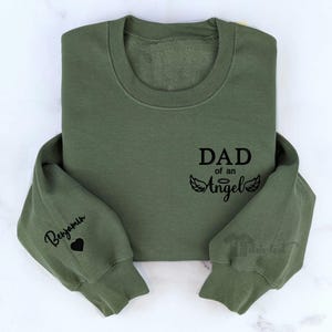 May include: Green sweatshirt with the text "DAD of an Angel" and a pair of angel wings. The left sleeve has the name "Benjamin" and a heart.
