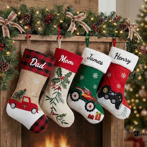 May include: Four Christmas stockings hang from a wooden mantel, each with a unique design. One is tan with a red truck, another is cream with holly, a green one with a monster truck, and a red one with a video game controller. The stockings are personalized with names.