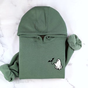May include: A sage green hooded sweatshirt featuring a small embroidered graphic on the chest. The design depicts a white ghost holding a black bat on a string. The hoodie has long sleeves with ribbed cuffs and a matching hood with a drawstring.