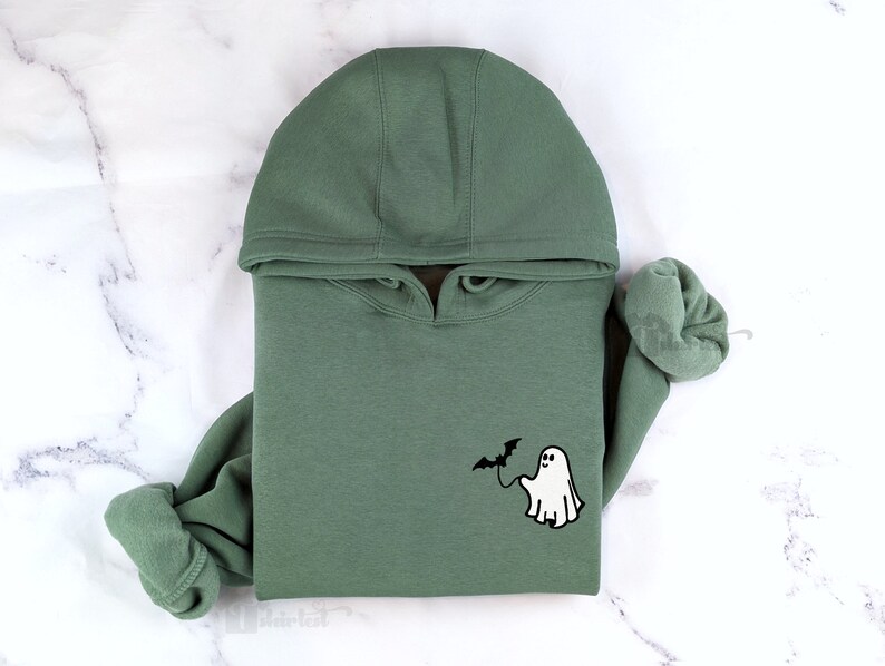 May include: A sage green hooded sweatshirt featuring a small embroidered graphic on the chest. The design depicts a white ghost holding a black bat on a string. The hoodie has long sleeves with ribbed cuffs and a matching hood with a drawstring.