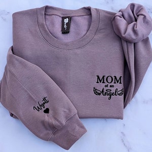 May include: A mauve sweatshirt with the text "MOM of an Angel" and angel wings printed on the front. The left sleeve has the name "Wyatt" and a heart printed on it.