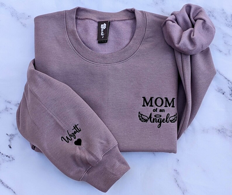 May include: A mauve sweatshirt with the text "MOM of an Angel" and angel wings printed on the front. The left sleeve has the name "Wyatt" and a heart printed on it.
