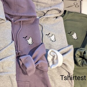 May include: Three hooded sweatshirts in purple, light gray, and olive green, each featuring an embroidered design of a white ghost holding a black bat on a string. The text "Tshirtest" is visible in the bottom right corner of the image.