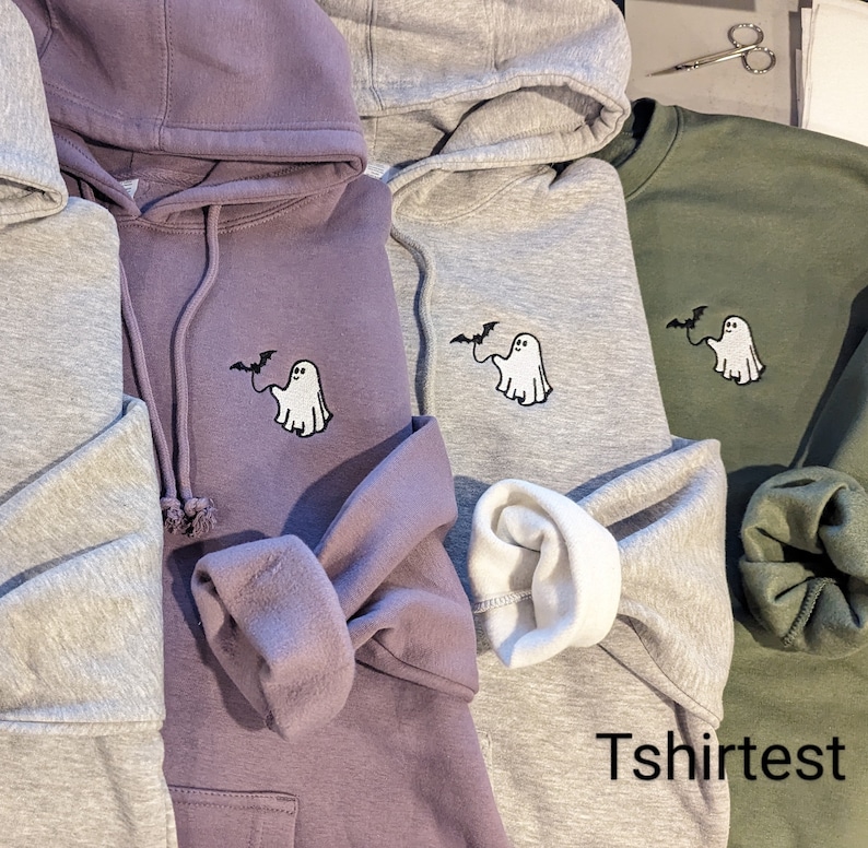 May include: Three hooded sweatshirts in purple, light gray, and olive green, each featuring an embroidered design of a white ghost holding a black bat on a string. The text "Tshirtest" is visible in the bottom right corner of the image.