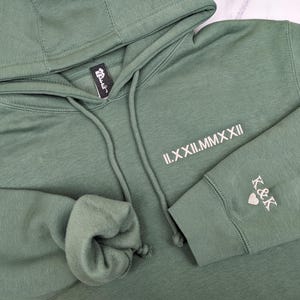 A thoughtful, custom anniversary gift. Detailed flat lay of a Sage Green hoodie featuring minimalist tusk Beige Roman numeral date embroidery on the chest and 'K&K' with a heart on the sleeve cuff