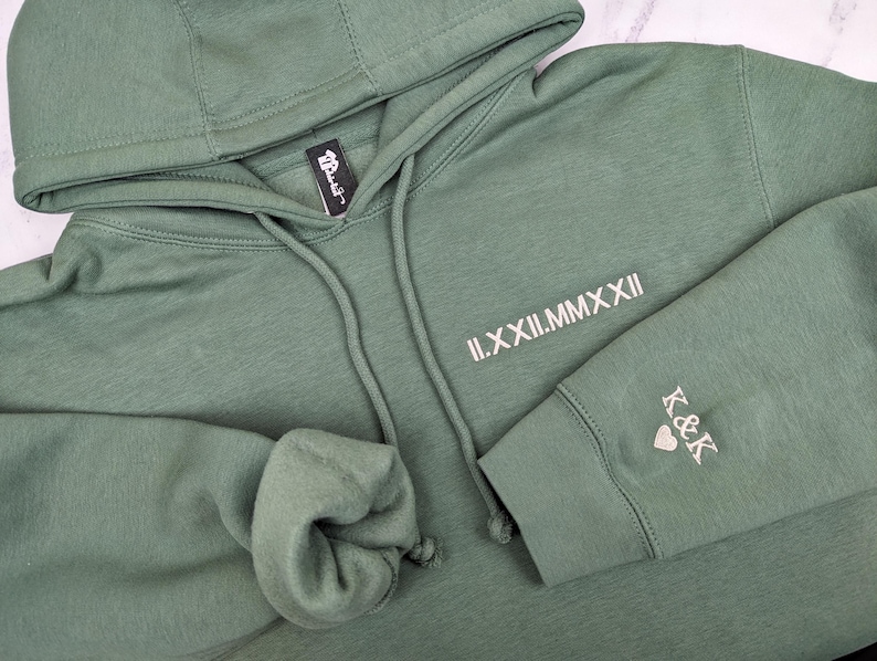 A thoughtful, custom anniversary gift. Detailed flat lay of a Sage Green hoodie featuring minimalist tusk Beige Roman numeral date embroidery on the chest and 'K&K' with a heart on the sleeve cuff