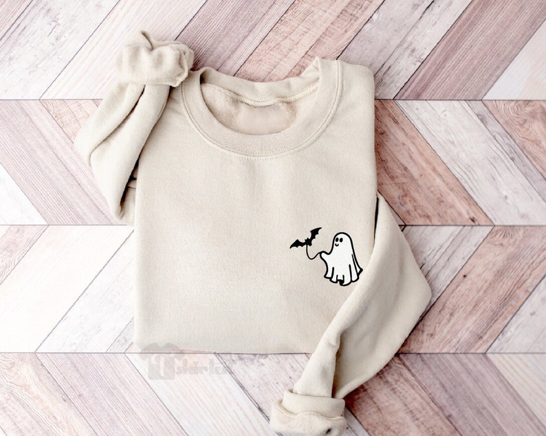 May include: A beige crewneck sweatshirt featuring a small black graphic on the left chest area. The graphic depicts a simple line-art ghost holding a string attached to a small flying bat. The sweatshirt has long sleeves with ribbed cuffs.