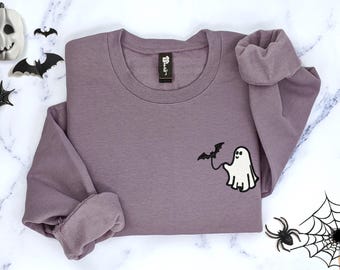 May include: A mauve long-sleeve crewneck sweatshirt featuring an embroidered design on the left chest. The embroidery depicts a small white ghost holding a string attached to a black flying bat. The sweatshirt has a ribbed collar and cuffs.