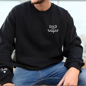 May include: Black sweatshirt with white text that reads "DAD of an Angel" and a pair of angel wings. The left sleeve has the name "Charlotte" and a heart.