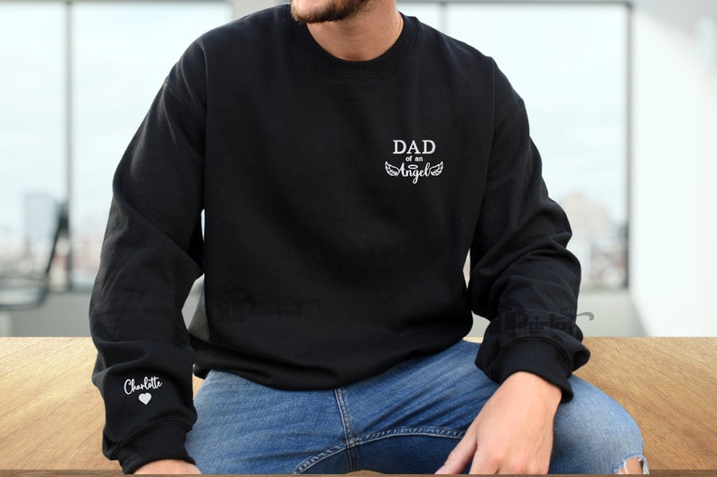 May include: Black sweatshirt with white text that reads "DAD of an Angel" and a pair of angel wings. The left sleeve has the name "Charlotte" and a heart.