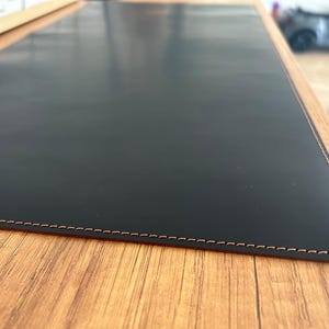 Puede incluir: Alfombrilla de escritorio de cuero negro con borde cosido. La alfombrilla está sobre una superficie de madera.