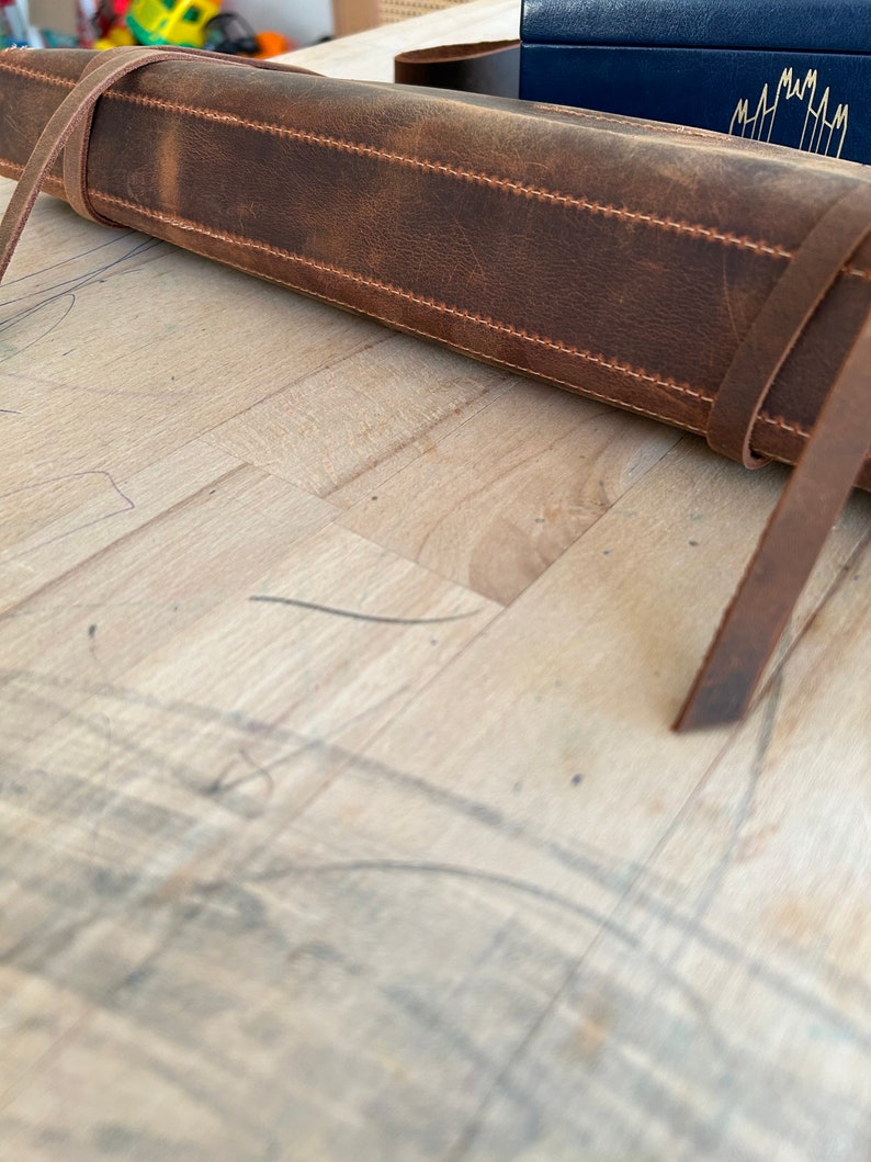 Op de afbeelding: Een bruine leren rolhoes met een band en stiksels. De hoes is opgerold en ligt op een houten oppervlak.