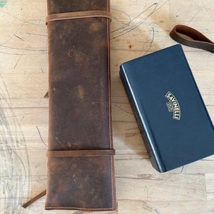 Op de afbeelding: Een bruine leren mesrol met twee banden en een blauwe doos met de tekst "Savinelli 1870" op de bovenkant.