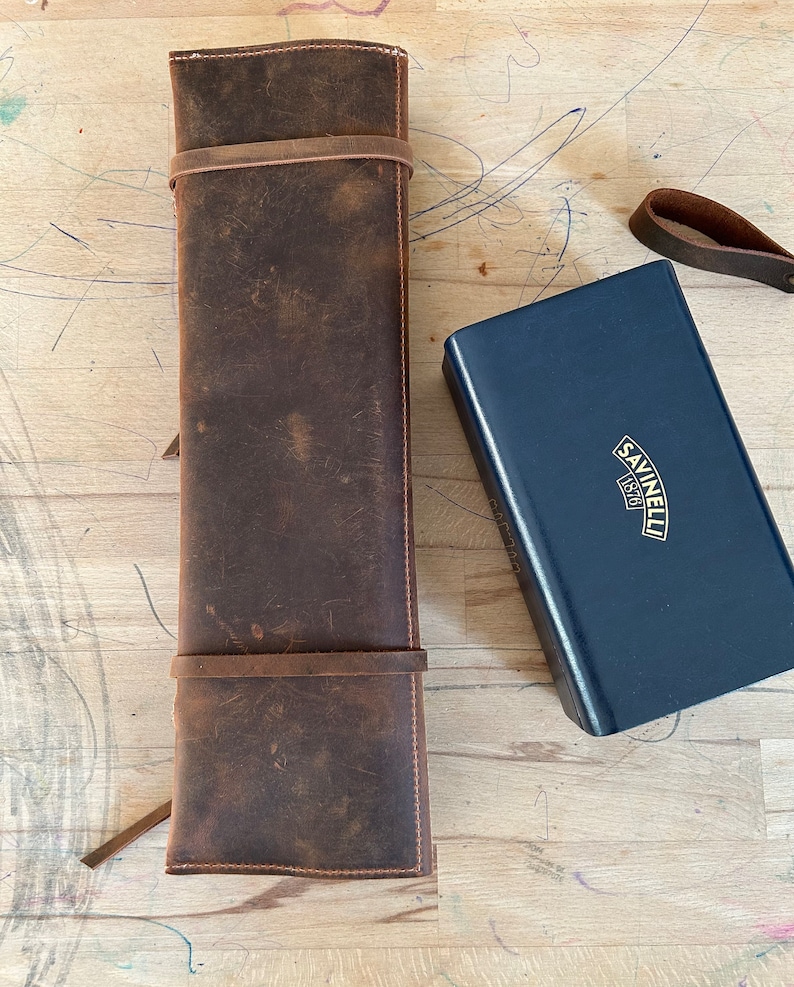 Op de afbeelding: Een bruine leren mesrol met twee banden en een blauwe doos met de tekst "Savinelli 1870" op de bovenkant.