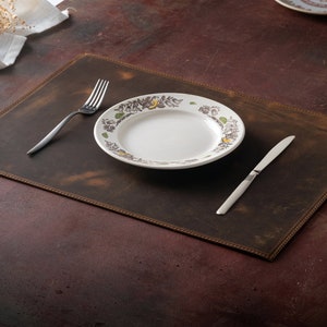 May include: A brown leather placemat with a stitched border, set with a white plate, fork, and knife. The plate has a decorative floral border. A wine glass is visible in the background.