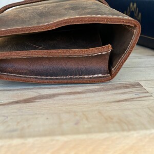Op de afbeelding: Een bruine leren tas met witte stiksels. De tas is dubbelgevouwen en de stiksels zijn zichtbaar aan de rand.