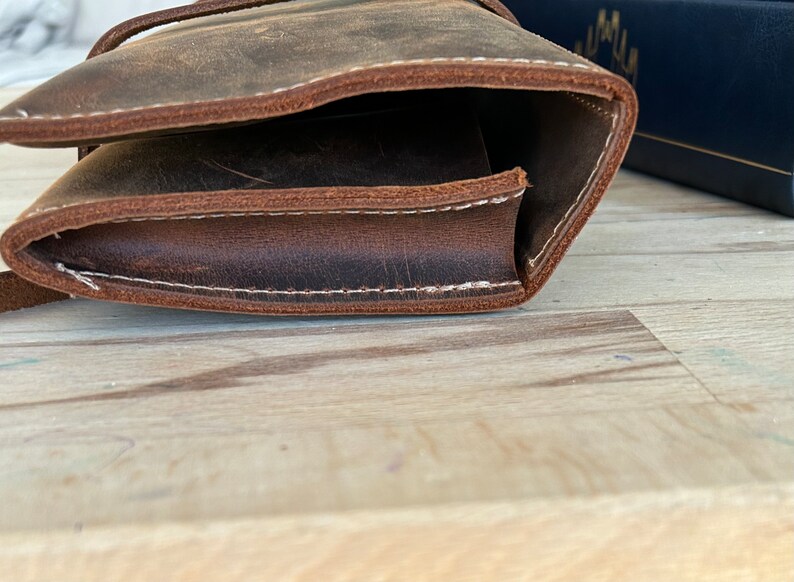 Op de afbeelding: Een bruine leren tas met witte stiksels. De tas is dubbelgevouwen en de stiksels zijn zichtbaar aan de rand.