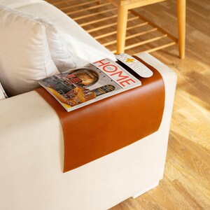 Könnte beinhalten: Ein braunes Leder-Armlehnen-Tablett mit einem Magazin und einer Fernbedienung darauf. Das Tablett ist an einem weißen Sofa befestigt.