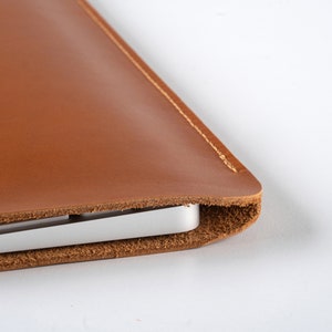 Puede incluir: Una funda de cuero marrón para portátil con un portátil plateado parcialmente insertado. La funda está hecha de cuero liso de color marrón y tiene una costura visible en el borde. El teclado del portátil es visible.