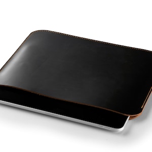 May include: A black leather tablet sleeve with a brown stitched edge. The sleeve is open and a tablet is partially visible inside.