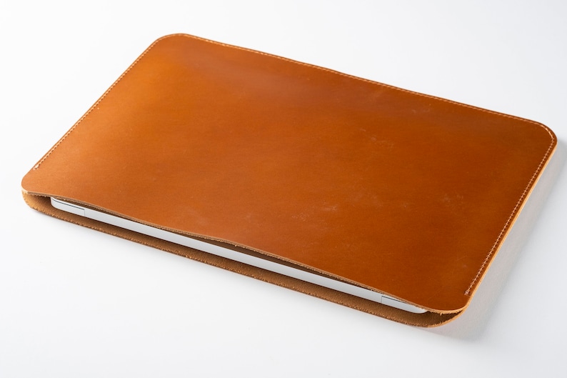 May include: A brown leather laptop sleeve with a subtle sheen. The sleeve is closed and the laptop is visible inside.