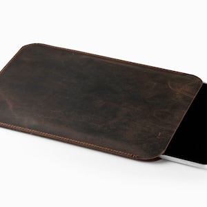 Può includere: Una custodia per tablet in pelle marrone con un bordo cucito. La custodia è aperta e un tablet nero è parzialmente visibile all'interno.